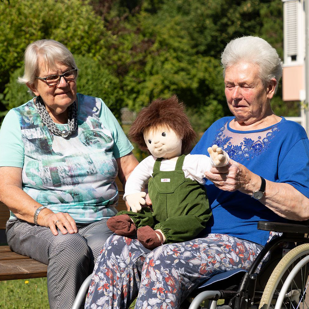 Two senior ladies engaging with the Joyk empathy doll Marco in the garden