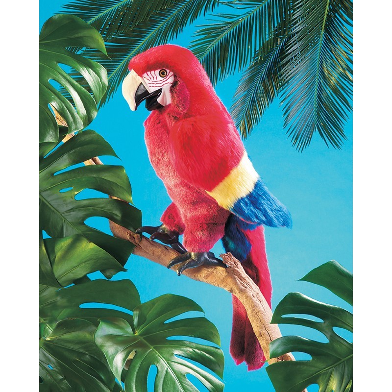 Folkmanis hand puppet scarlet macaw in vivid red with yellow-blue wings, perched on a branch against a turquoise-blue background with monstera leaves