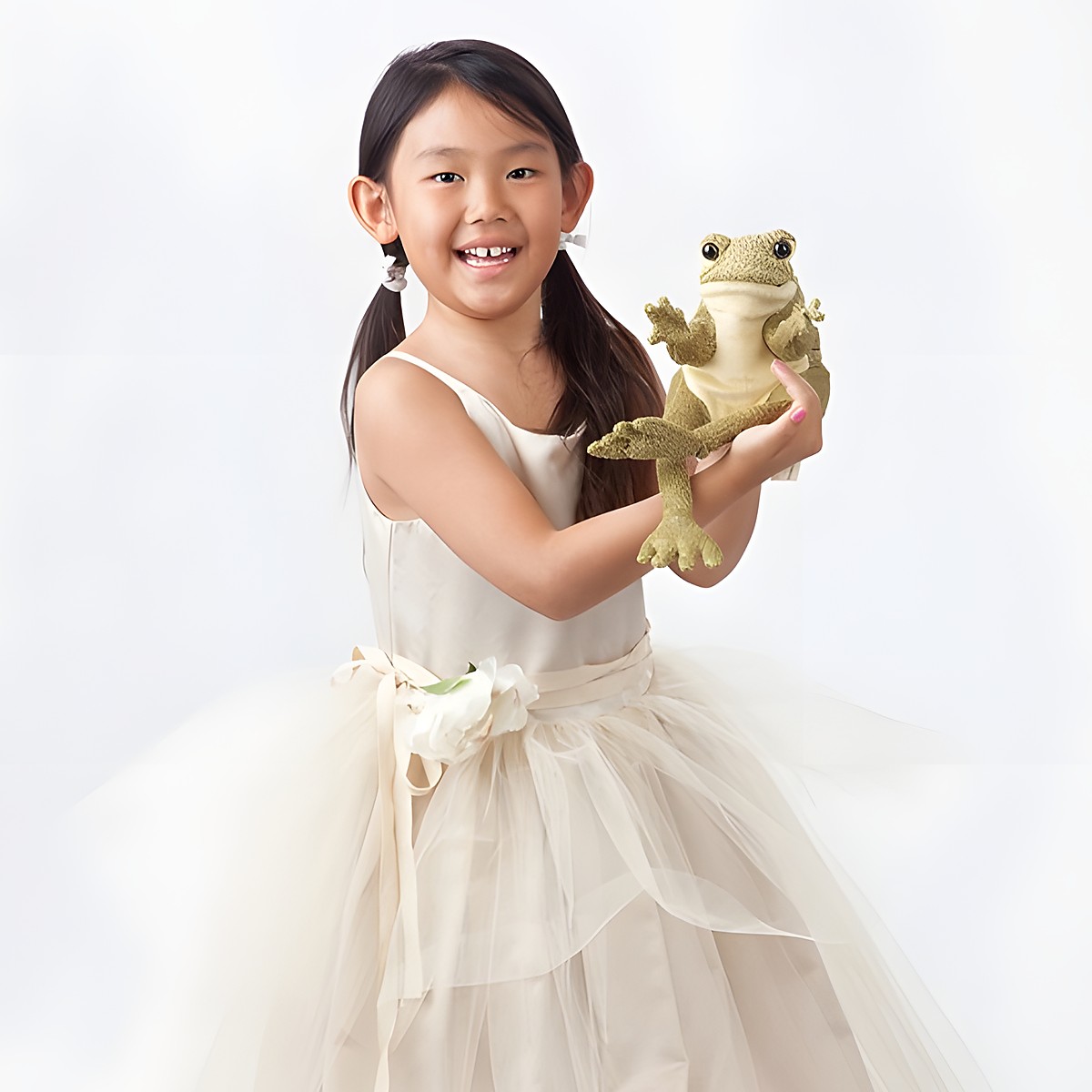 Girl in a cream tulle dress holding the Folkmanis Funny Frog hand puppet in olive-green plush up to the camera