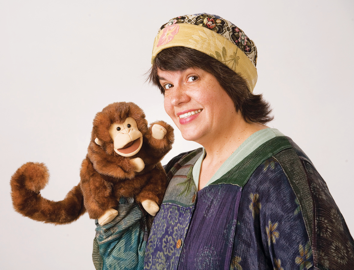 Folkmanis monkey hand puppet with brown fur and movable mouth, operated by a woman wearing a hat, held over her shoulder