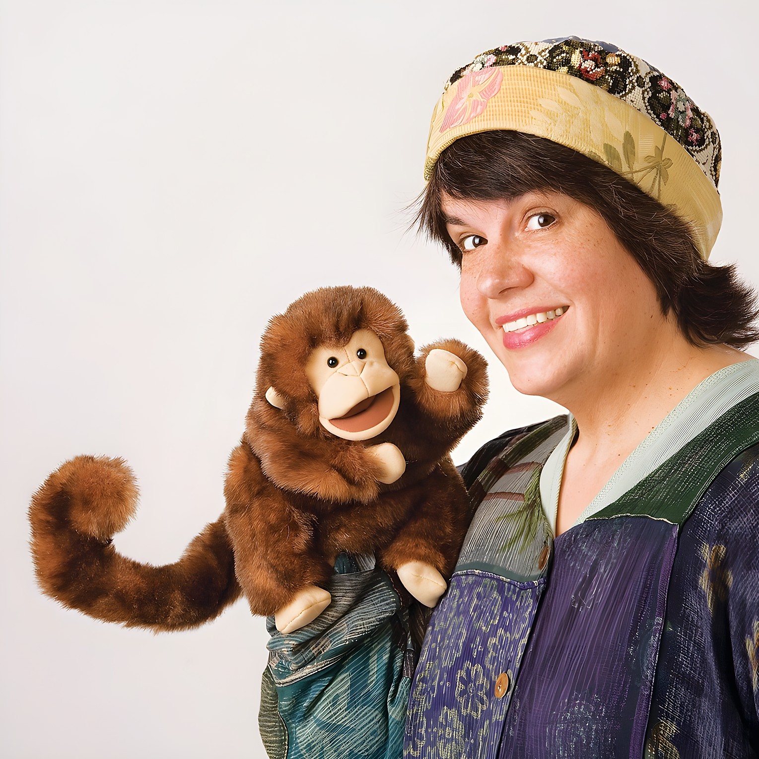 Folkmanis monkey hand puppet with brown fur and movable mouth, operated by a woman wearing a hat, held over her shoulder