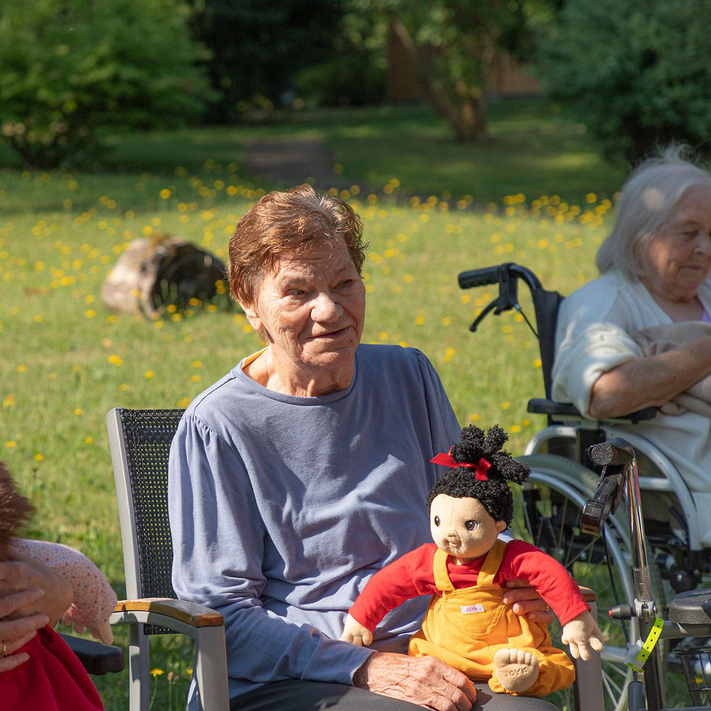 Senior lady with the Joyk empathy doll Naomi on her lap during a garden gathering