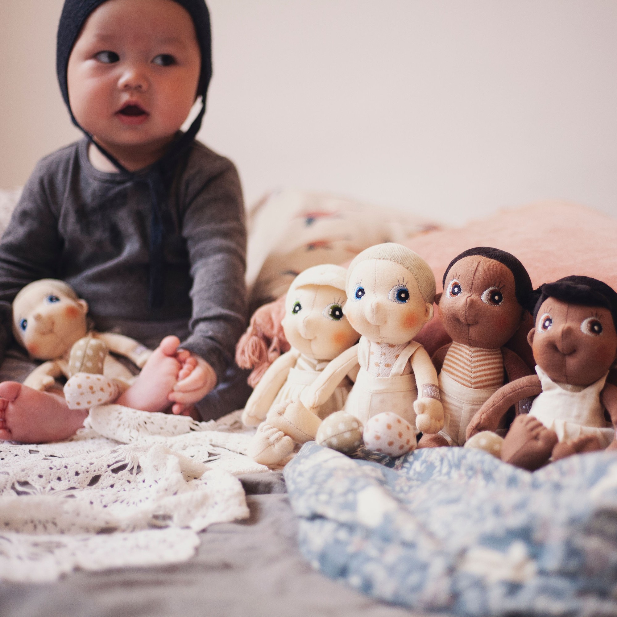 Baby sitting next to a row of Rubens EcoBuds mini dolls on the bed