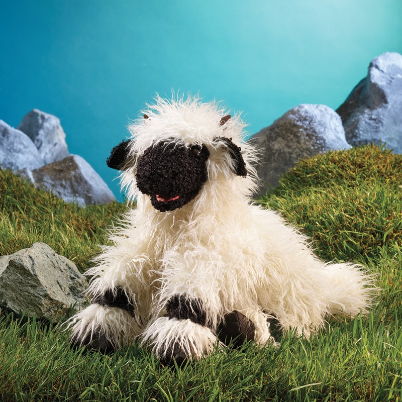 Folkmanis hand puppet Valais Blacknose Sheep with white shaggy fur and black nose, lying on grass in front of a rocky backdrop