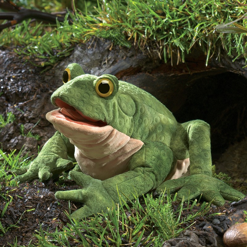 Folkmanis toad hand puppet in rich green with textured skin, open mouth and yellow eyes, resting on damp ground in front of a tree hollow