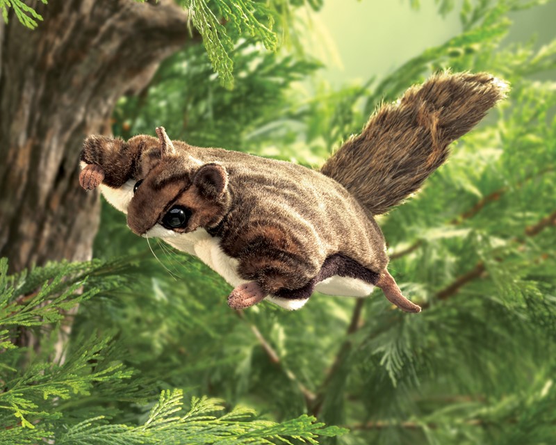 Folkmanis flying squirrel hand puppet in brown plush, photographed in gliding pose among green fir branches