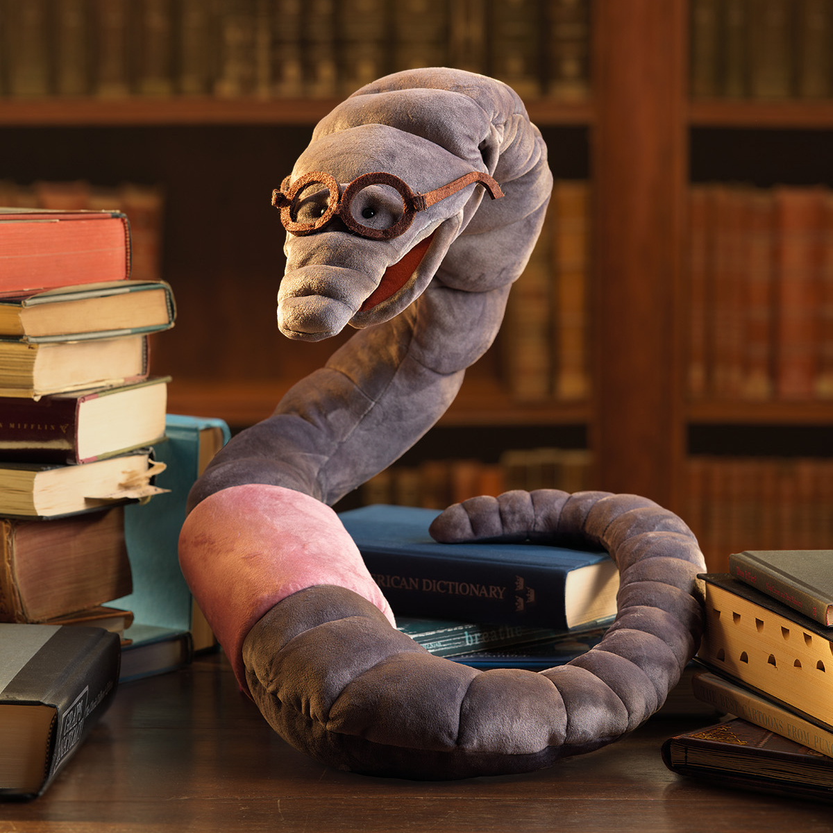 Folkmanis bookworm hand puppet in grey and pink plush with round glasses and open mouth, rearing up between stacks of books in a library setting