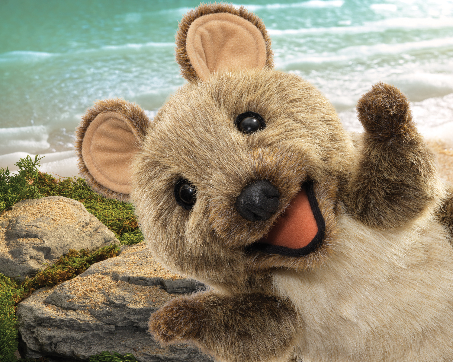 Folkmanis quokka hand puppet in brown plush with open mouth and raised arm, posed against a beach backdrop