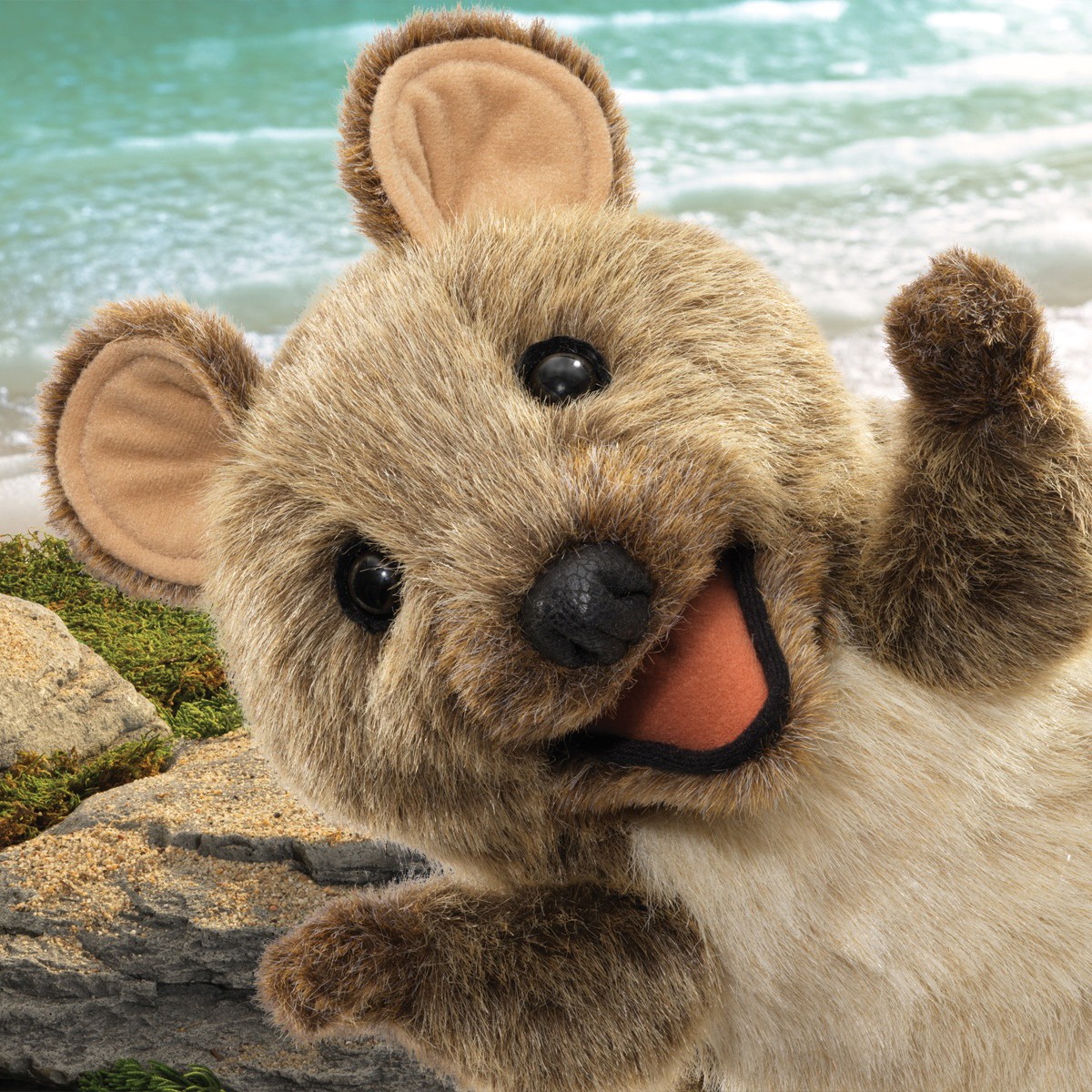 Folkmanis quokka hand puppet in brown plush with open mouth and raised arm, posed against a beach backdrop