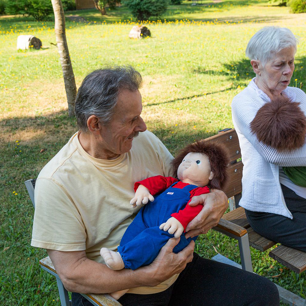 Senior gentleman holding the Joyk empathy doll Johan in his arms during a garden gathering