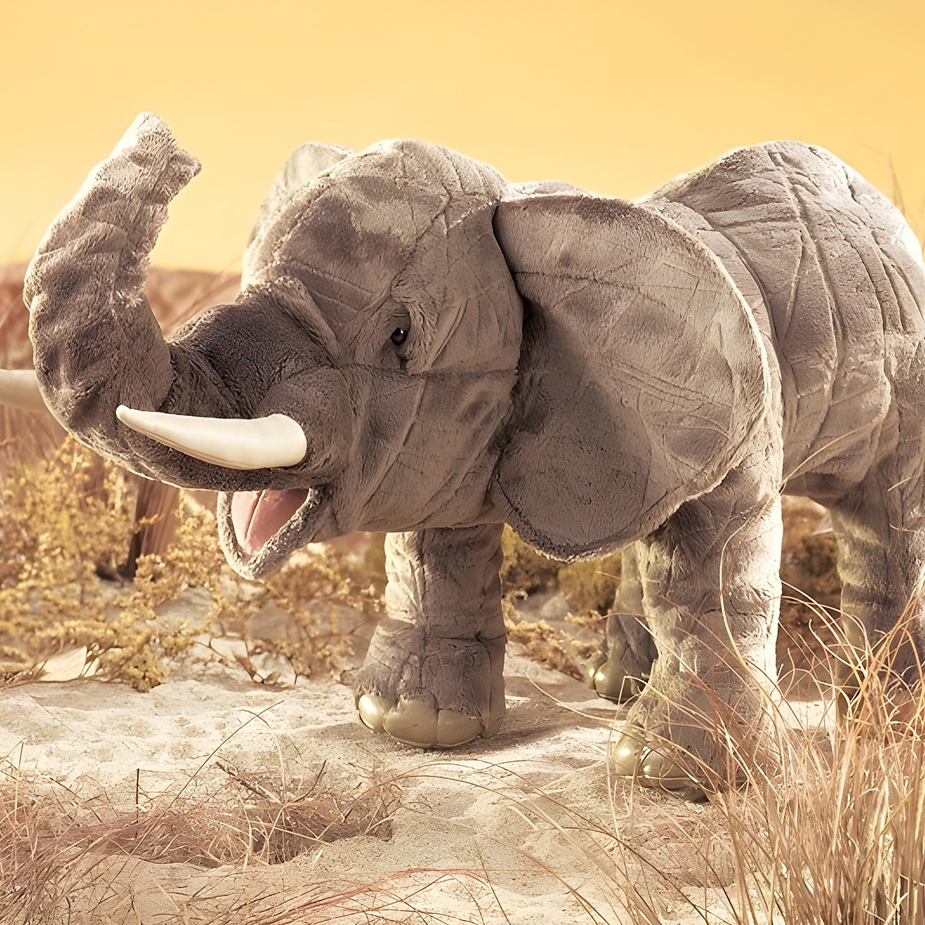Folkmanis elephant hand puppet in grey plush with raised trunk, open mouth and white tusk, standing in front of a savannah backdrop