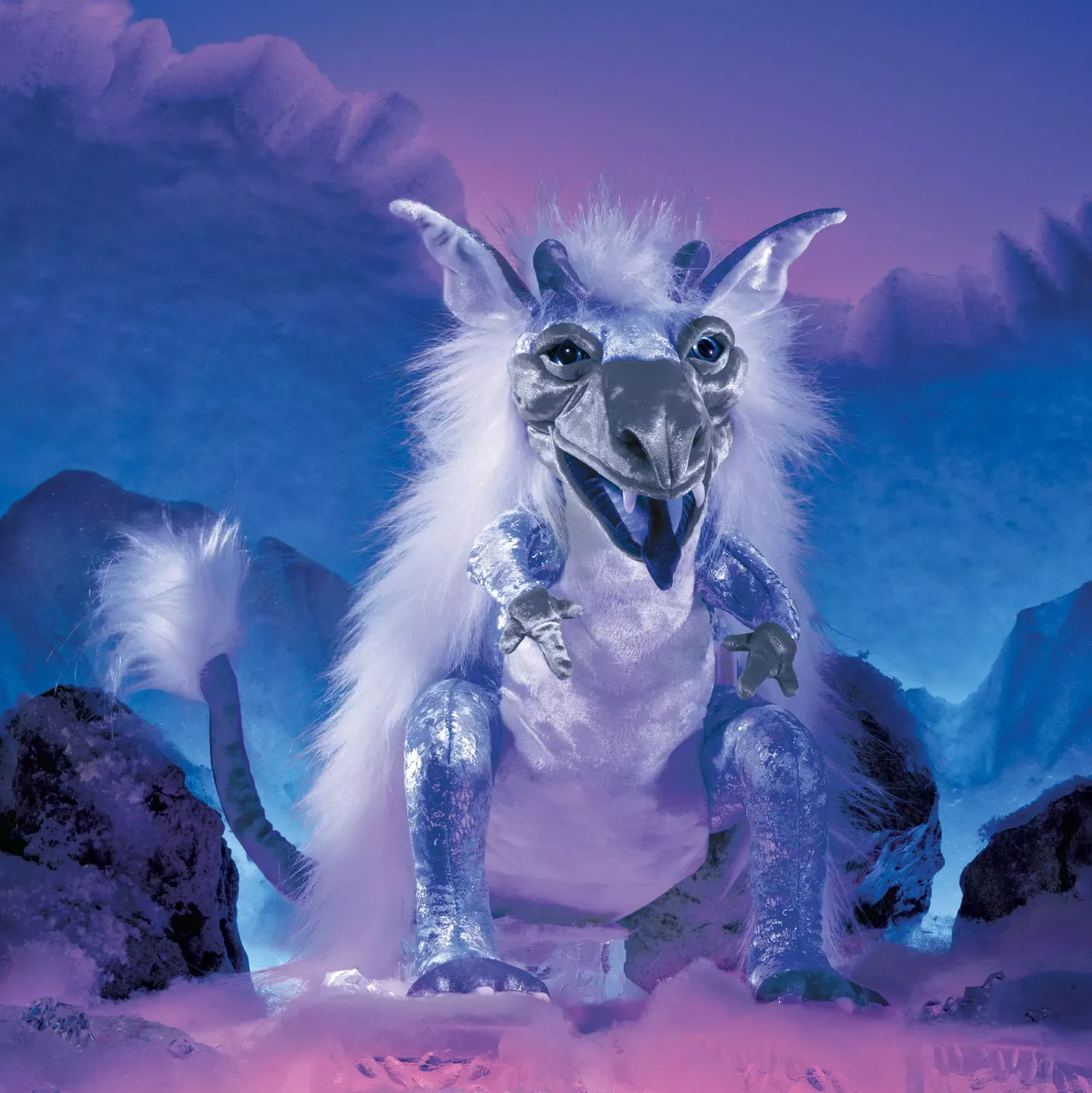 Folkmanis hand puppet ice dragon in silver-white with open mouth, horns and fluffy mane against an icy blue mountain backdrop