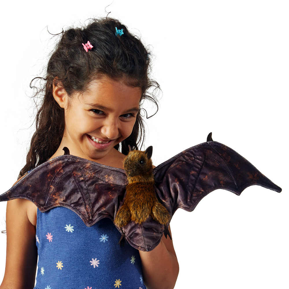 Girl holding a Folkmanis fruit bat hand puppet with spread dark brown wings and a rust-brown plush body toward the camera