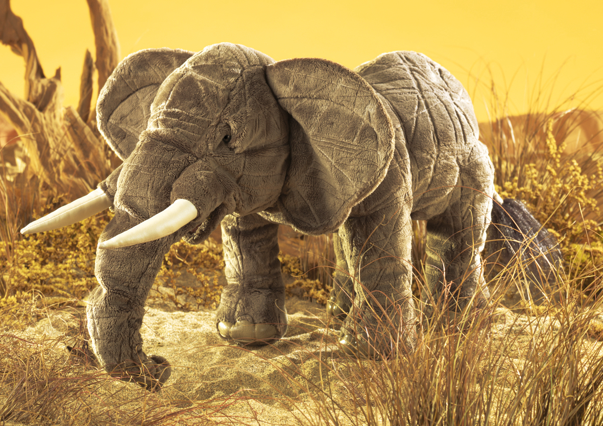 Folkmanis hand puppet elephant in grey with white tusks, trunk pointing down, side view against a yellow savanna background