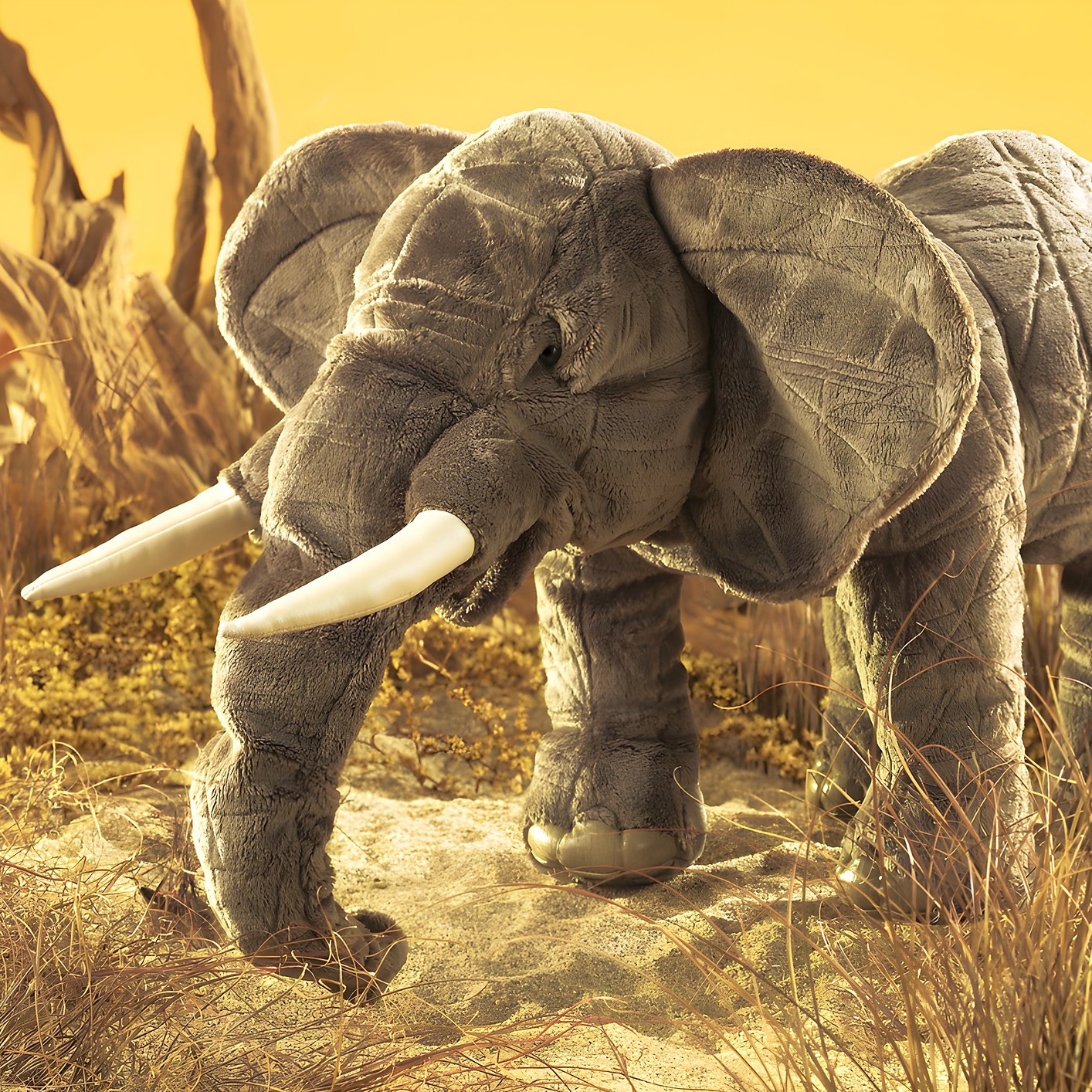 Folkmanis hand puppet elephant in grey with white tusks, trunk pointing down, side view against a yellow savanna background