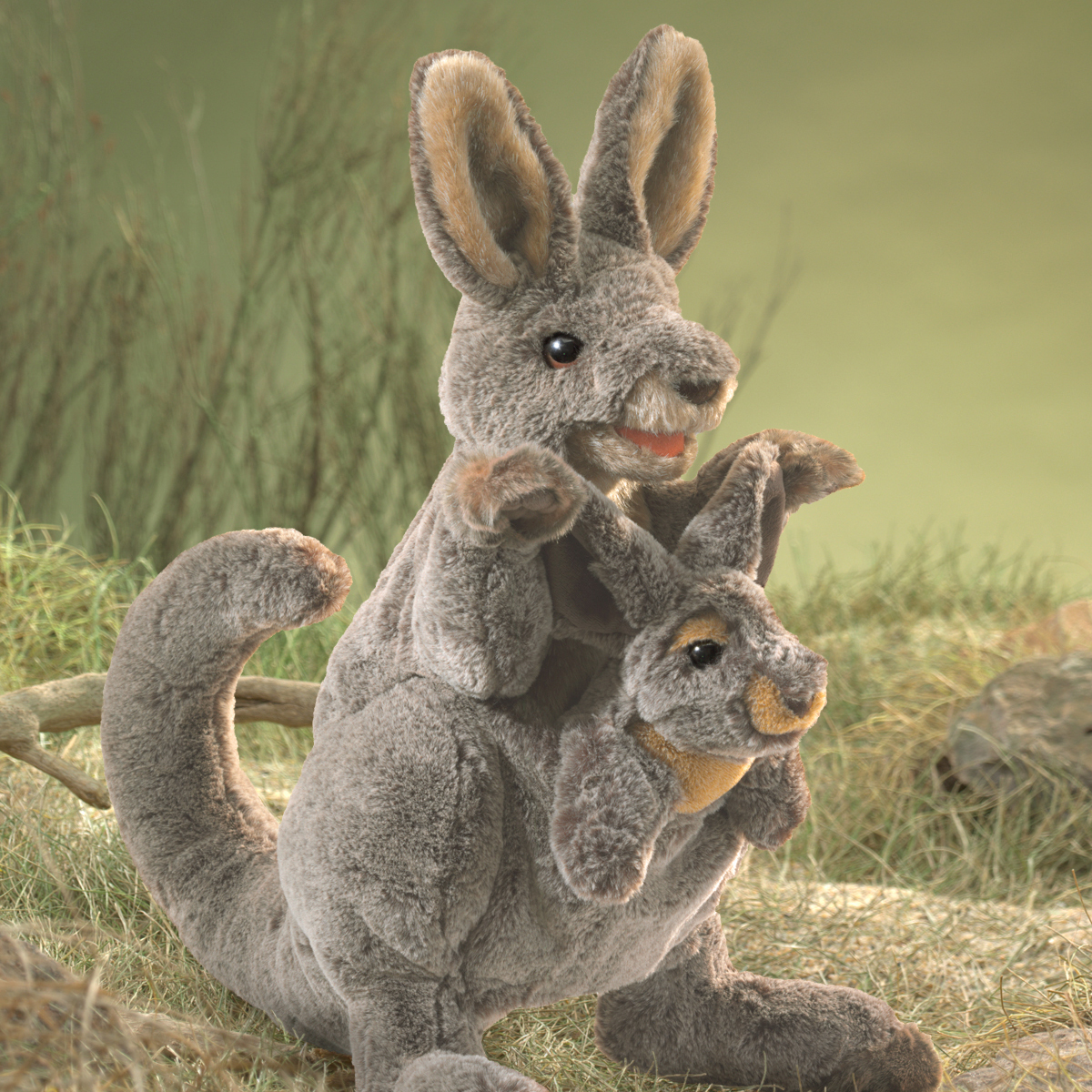 Folkmanis hand puppet kangaroo with baby in grey plush, joey peeping out of the pouch, front view against a grassland backdrop