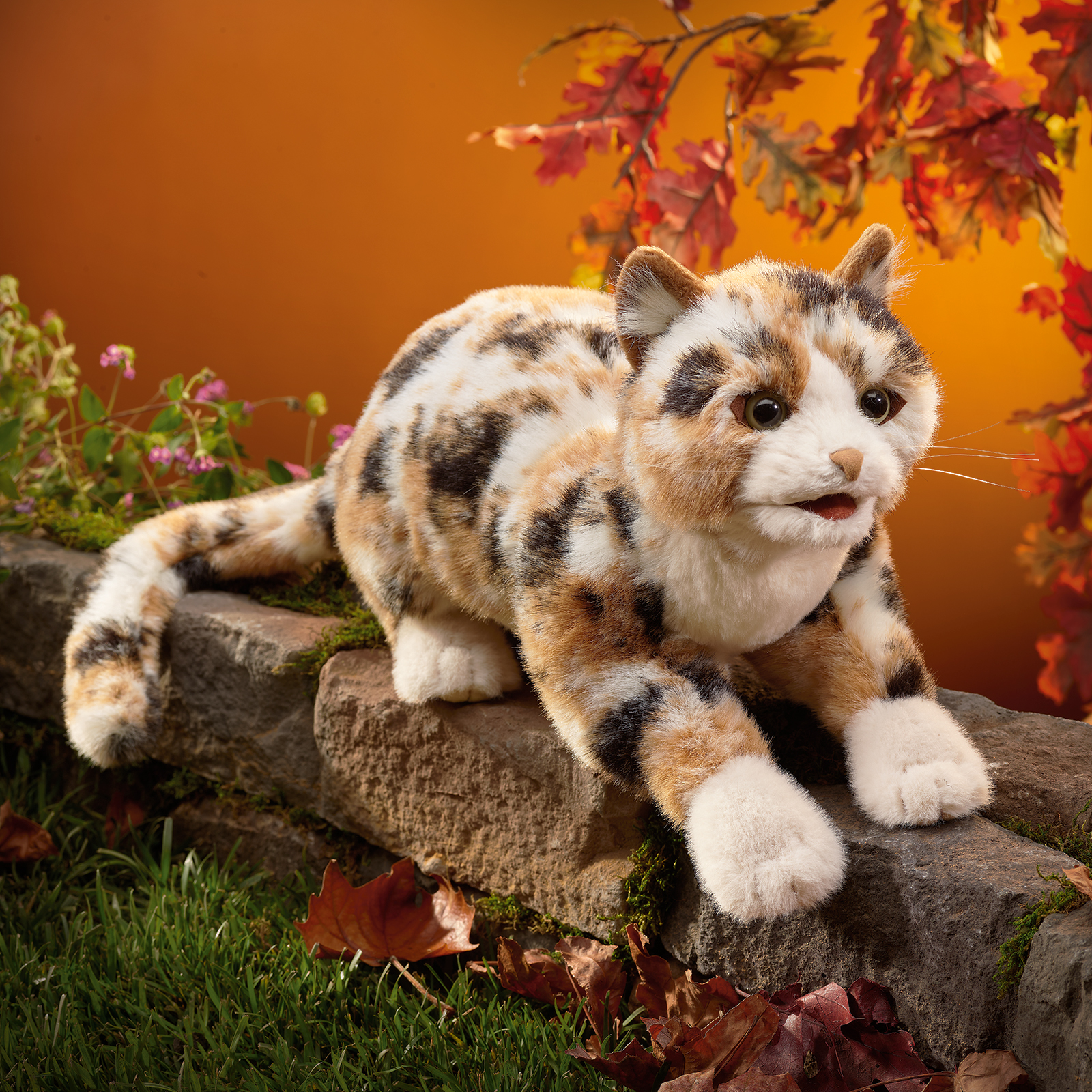 Folkmanis calico cat hand puppet with tricolour fur in white, orange and black, lying on a stone wall, with a movable mouth