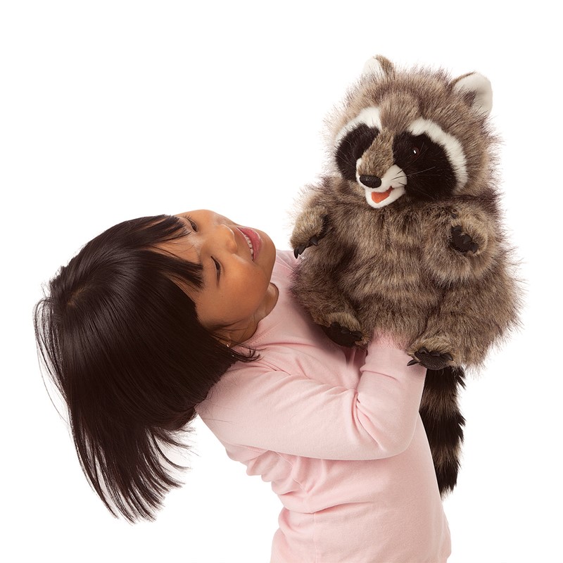 Girl holding a Folkmanis raccoon hand puppet with an open mouth up in the air, grey-brown fur with a black face mask