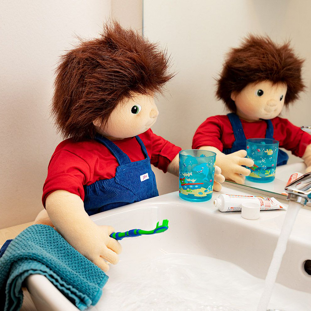 The Joyk empathy doll Johan at the washbasin with a toothbrush cup in front of the mirror