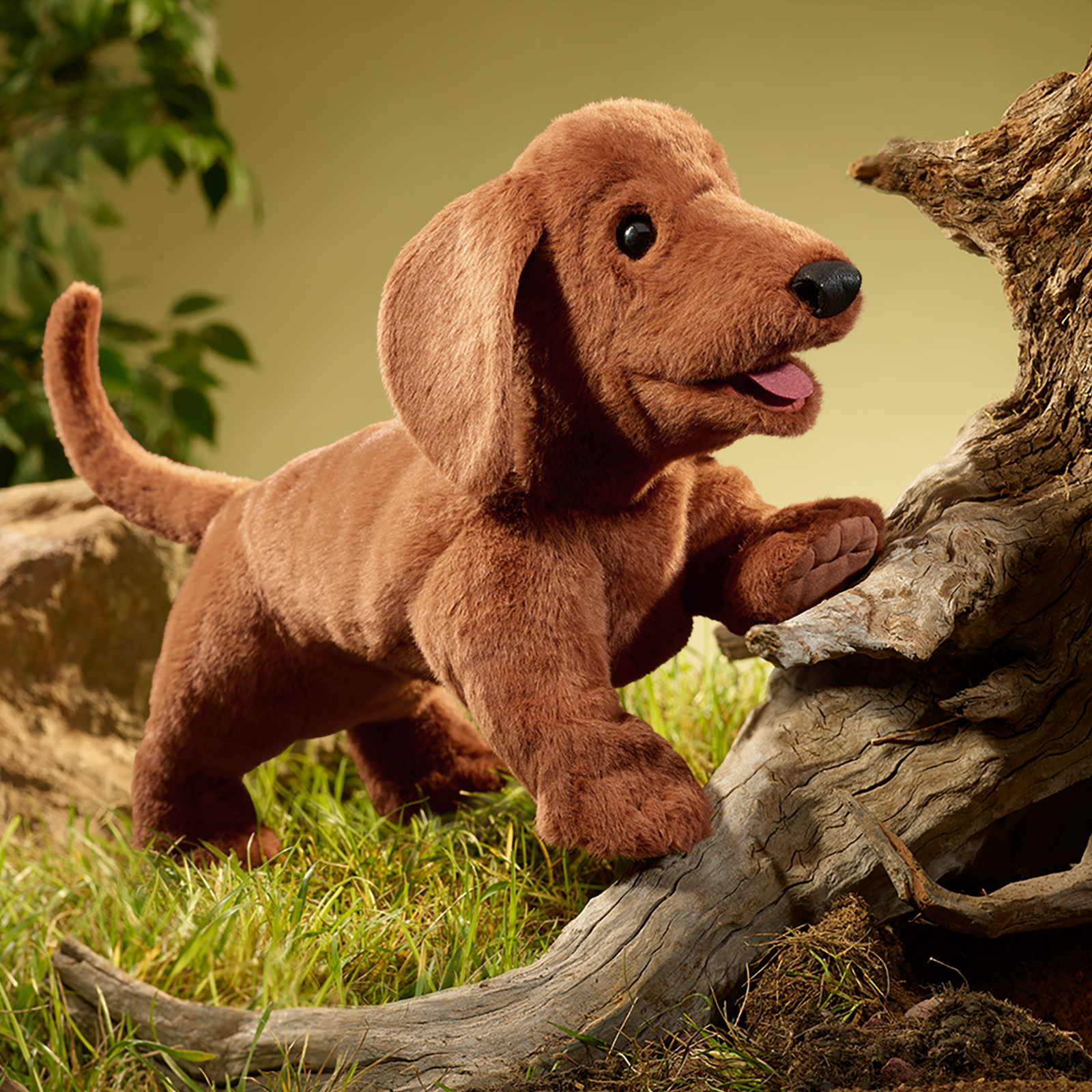 Folkmanis dachshund hand puppet resting one front paw on a branch, mouth open, with grass and leaves behind it.