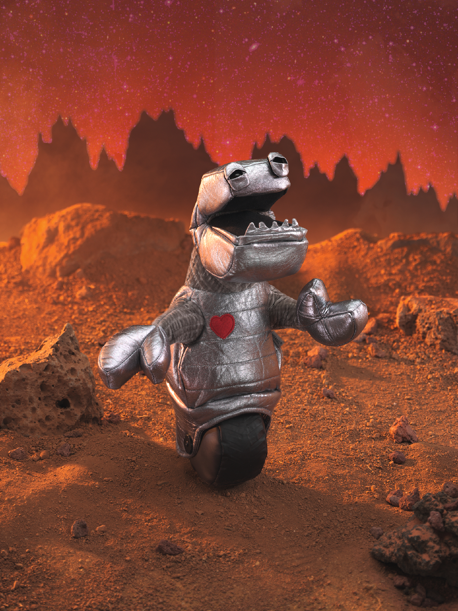 Folkmanis robot hand puppet in a silver full-body suit with a red heart on the chest, open mouth and outstretched claw hand, against a Mars-like backdrop