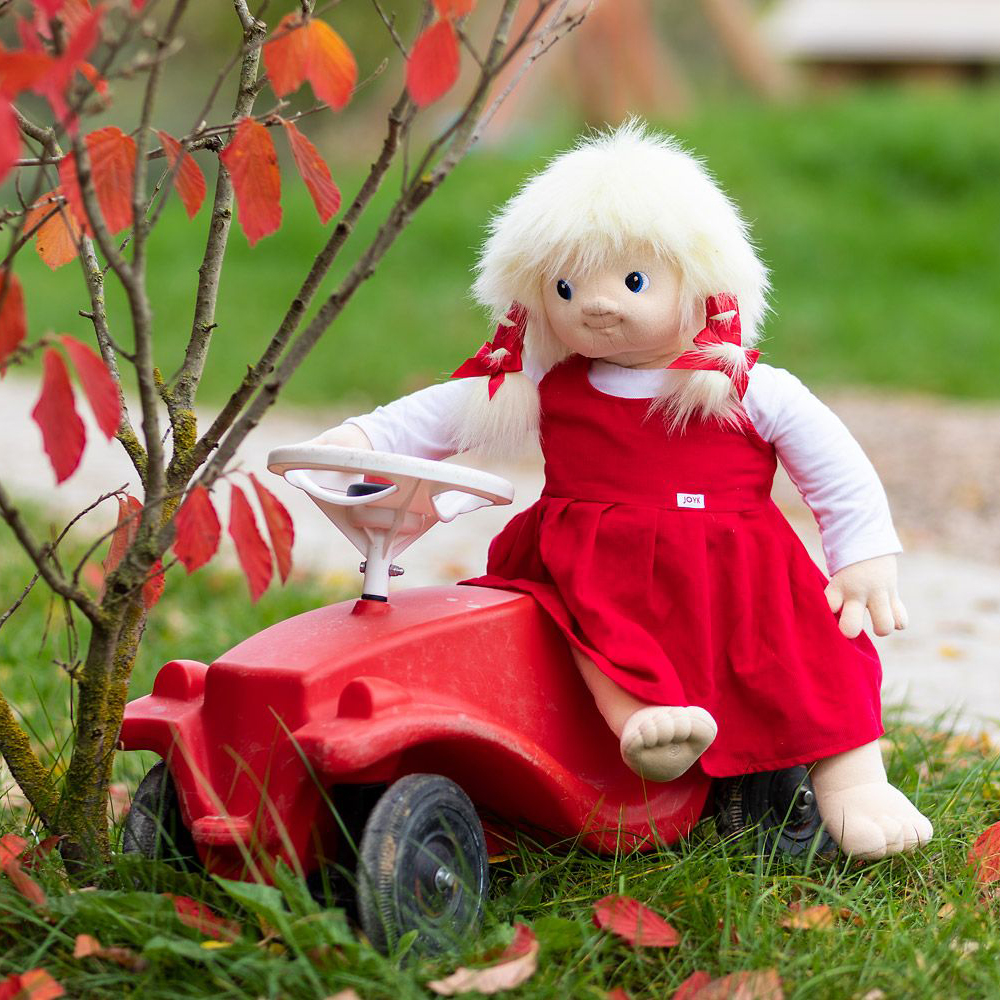 The Joyk empathy doll Nelly sitting on a red ride-on car in an autumn garden