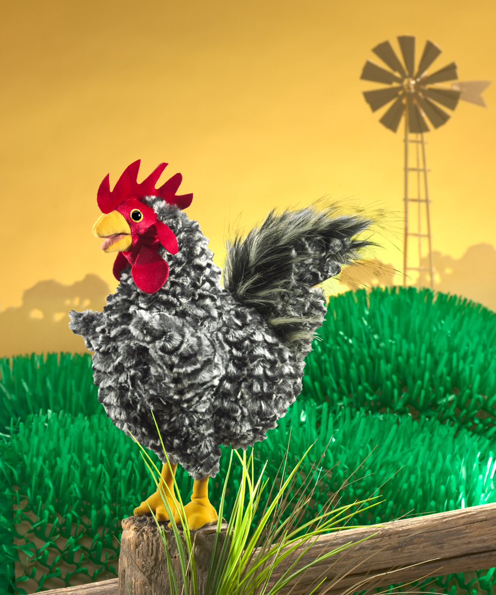 Folkmanis hand puppet rooster with grey-white curly plumage, red comb and yellow claws, perched on a wooden post