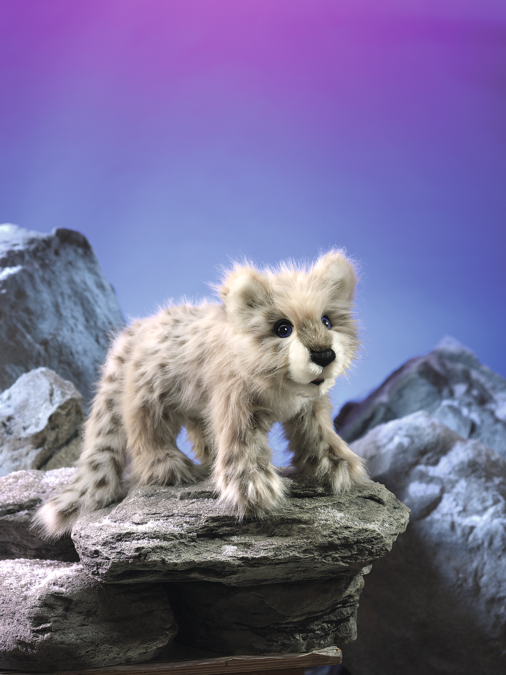 Folkmanis hand puppet baby snow leopard with grey-beige spotted fur, standing on a rock against a blue-violet background